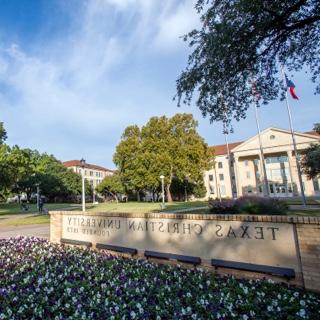 A brick sign bearing the name Texas Christian University sits behind a flowerbed of purple pansies